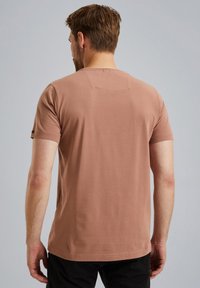 Krátký rukáv, hnědé bavlněné tričko s kulatým výstřihem; má minimalistický design a hladkou texturu. Na zadní straně se nachází malý horizontální šev.