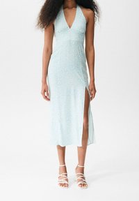 Vaaleansininen kukkakuosinen mekko, jossa on halterneck-kaula-aukko, reidet paljastava halkio ja sileä kangas; yhdistetty valkoisiin remmikkäisiin korkokenkiin.