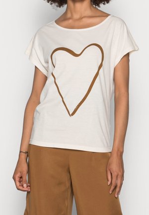 Frau trägt ein weißes, locker sitzendes T-Shirt mit einer großen braunen Herzumrandung, kombiniert mit braunen Hosen vor einfarbigem Hintergrund.