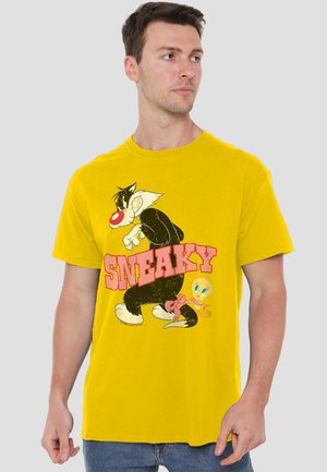 Young man wearing a yellow T-shirt with a cartoon cat and bird facing opposite directions and the word "SNEAKY" in bold pink letters.