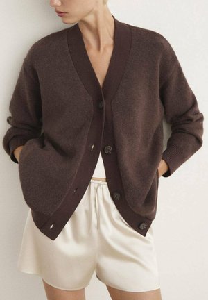 Cardigan en laine marron avec un col en V, des boutons sur le devant et des poignets côtelés. Porté sur un haut blanc et un short crème. Texture lisse.