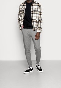 Camisa de cuadros de botones en blanco, negro y dorado sobre un cuello redondo negro. Pantalones de chándal gris ajustados y zapatillas negras con suelas y cordones blancos.