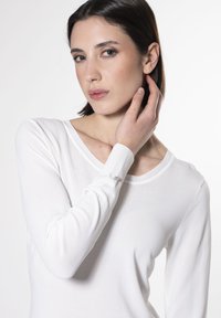 Top bianco a maniche lunghe con scollo rotondo e polsini a costine. Presenta una texture liscia e una leggera lucentezza sull'accento dei polsini.