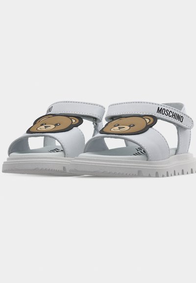 Λευκά σανδάλια παιδικά με καφέ πρόσωπα αρκουδάκια στα μπροστινά λουριά και λουριά στον αστράγαλο με την επωνυμία "Moschino" σε λευκό φόντο.