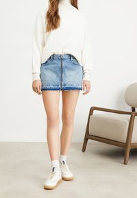 Gonna mini in denim azzurro chiaro con orlo sfilacciato, dotata di zip frontale e tasche laterali. Abbinata a un maglione crema e sneakers bianche.