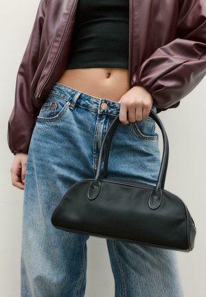Osoba v bordové bundě a černém krátkém topu držící černou koženou kabelku, doplněnou o vysokým pasem modré džíny.