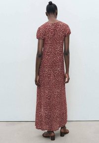 Robe maxi bordeaux avec des motifs floraux blancs et beiges, manches courtes bouffantes, col rond et design fluide. Portée avec des sandales brunes.