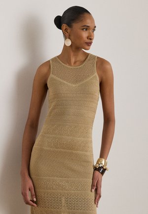 Nainen, jolla on tumma nuttura, pukeutuneena hihattomaan beigen sävyiseen neulemekkoon, suuret pyöreät korvakorut ja useita rannekoruja ranteessaan.