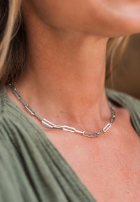 Necklace u obliku srebrne spajalice s izduženim, međusobno povezanim segmentima. Glatka, polirana tekstura na pozadini zelene tkanine.