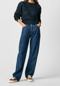 Marineblauwe gebreide trui met een gestructureerd patroon en ronde hals, gecombineerd met hoogwater, losvallende blauwe jeans en bruine sneakers.