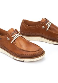 Chaussures décontractées marron en cuir à enfiler avec lacets élastiques blancs et semelles en caoutchouc beige clair tachetées sur fond blanc.