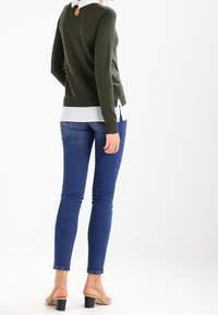 Olivgrön tröja med nyckelhålsrygg, lager på en vit skjorta. Kombinerad med blå skinny-jeans och beige blockklacksandaler.