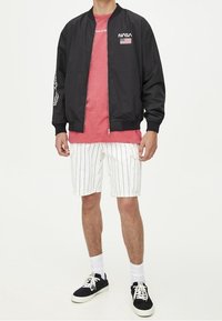 Sort bomberjakke med NASA-logo, lyserød T-shirt, hvide stribede shorts og sorte sneakers med hvide såler. Model står mod en lys baggrund.