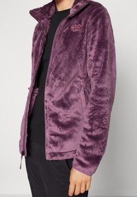 Person som bär en lila plysch zip-up-jacka över en svart skjorta med synliga dragkedje-fickor på jackans framsida.