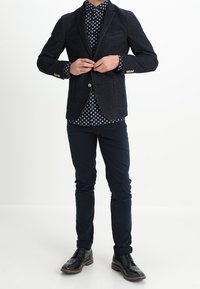 Marineblazer met twee zakken, gestructureerde stof, koperen knopen. Daaronder een donkerblauw polkadot overhemd, gecombineerd met slimfit marineblauwe broek en zwarte schoenen.