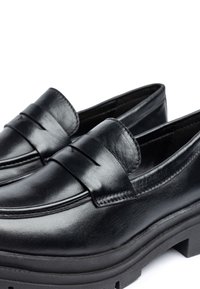Mocasines de cuero negro con suela plana y gruesa, y una tira frontal detallada con pliegue. Textura suave con costuras limpias y un diseño minimalista.