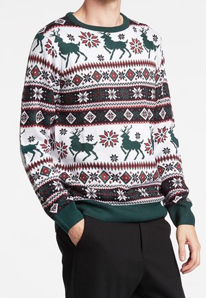 Maglione festivo con renne verdi, fiocchi di neve rossi e bianchi e motivi geometrici su sfondo bianco, indossato con pantaloni neri.
