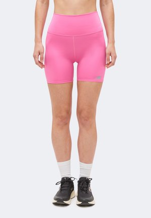 Žena u jarkoružičastim sportskim kratkim hlačama s visokim strukom, bijelim sportskim čarapama i crnim tenisicama za trčanje stoji ispred jednobojne pozadine.