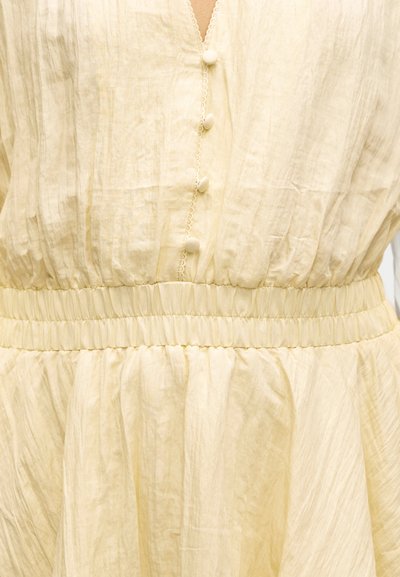 Μαλακό κίτρινο φόρεμα με σούρες στη μέση, ντεκολτέ σε σχήμα V και μικρά κουμπιά καλυμμένα με ύφασμα κατά μήκος του μπροστινού μέρους.