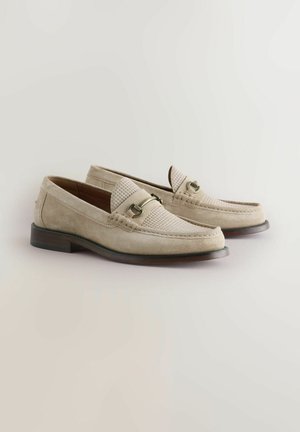 Beige Wildleder-Slipper mit strukturierten Akzenten, abgerundeter Zehenpartie, dekorativem Riemen und niedrigem Holzabsatz. Glatte Oberfläche mit Nahtdetails.