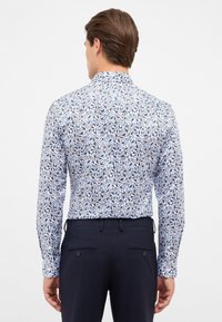 Wit longsleeve shirt met blauwe bloemenpatronen, voorzien van een gestructureerde kraag en knoopmanchetten. Draagt met marineblauwe broek.