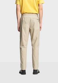 Homme portant un pantalon décontracté beige avec une ceinture élastique et une poche zippée à l'arrière, associé à des chaussures noires et une chemise jaune, debout tourné vers l'arrière.
