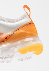 VaporMax-sneakers med en vit och orange ovandel, sömlös design, genomskinlig sula och en strukturerad hälflik för grepp och stöd.