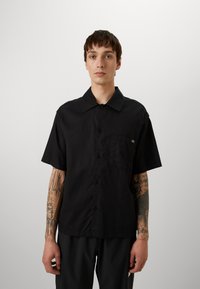 Black short-sleeve button-up shirt with a front pocket, made of a lightweight fabric. Simple design with a pointed collar and logo accent.