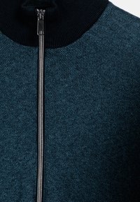 Gebreide zip-up jas in donkerblauwgroen met een gestructureerde afwerking, voorzien van een zwarte geribde kraag en een zilveren rits aan de voorkant.