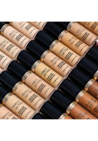 Collection of concealer bottles arranged in rows. Bottles feature a gold-capped design, various skin tones, and labeled "Born This Way."