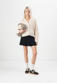 Beige geribbelde cardigan met twee knopen, zwarte plooienrok en zwarte sneakers met witte accenten. Houdt een tas met slangenhuidpatroon vast.