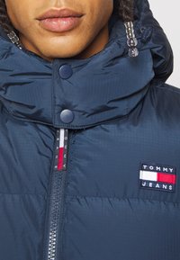 Granatowa puchowa kurtka z wysokim kołnierzem, widocznym zamkiem błyskawicznym i zatrzaskami. Posiada naszywkę z logo Tommy Jeans oraz fakturowaną tkaninę.