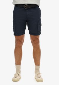 Pantalones cortos de carga de color navy, hechos de un tejido ligero, con dos bolsillos laterales y puños ajustables. Combinados con zapatillas beige y calcetines en tonos claros.