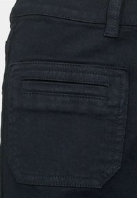 Melnas denima audums ar aizmugures kabatu ar horizontālu atvērumu. Auduma struktūra ir gluda ar maigu spīdumu un redzamām šuvju detaļām.