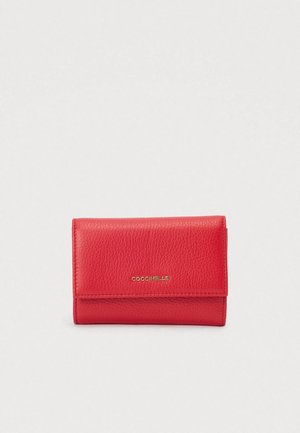 Červená texturovaná kožená peněženka složená a uzavřená se zlatým embosovaným logem "COCCINELLE" ve středu na přední straně na jednobarevném pozadí.