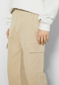 Beżowe spodnie typu cargo wykonane z wytrzymałej tkaniny, z kieszeniami z przodu i po bokach, podkreślonymi przeszyciami i luźnym krojem.