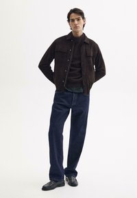 Braune Wildlederjacke mit zwei Brusttaschen, brauner Pullover darunter, blaue Jeans und schwarze Schuhe; Modell steht mit Händen in den Taschen.