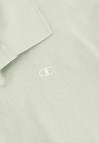 Hellgrünes Poloshirt aus Baumwollstoff, mit Kragen und subtil gesticktem Logo auf der Brust. Sanfte Textur, lässiges Design.