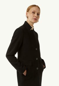 Schwarzer Wollmantel mit collarlosem Design, ausgestattet mit einer Knopfleiste und seitlichen Taschen. Der Stoff hat eine glatte Textur.