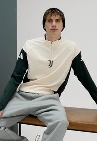Collared long-sleeve sports top in cream with dark green sleeves. Features a half-zip and a logo on the chest. Paired with light gray pants.