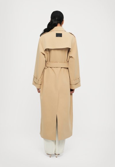 Μπεζ παλτό με ζώνη στη μέση, μακριά μανίκια και αεραγωγό στην πλάτη. Διαθέτει ένα ορθογώνιο λογότυπο στην άνω πλάτη.