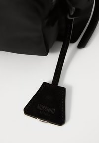 Čierna kožená kabelka s našitým štítkom Moschino Milano pripevneným tenkým popruhom na bielom povrchu.