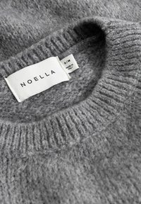 Pilkas megztos medžiagos megztinis su briaunuotu iškirptės kraštu ir etikete, kurioje užrašyta „NOELLA“. Dydžio žymė S/M, pažymėta „PAGAMINTA ITALIJOJE“.