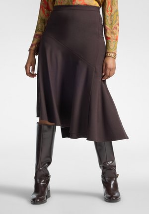 Femme portant une jupe asymétrique marron foncé, un haut à manches longues à motifs et des bottes brillantes marron foncé montant jusqu'aux genoux, debout avec les jambes écartées.