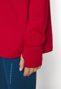 Main reposant sur le côté portant une chemise rouge à manches longues avec trou pour le pouce et un pantalon bleu avec des points blancs verticaux.