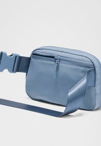 Bolso azul de textura suave con un compartimento principal con cremallera, una correa de nylon ajustable y una hebilla de plástico resistente. Diseño suave y minimalista.