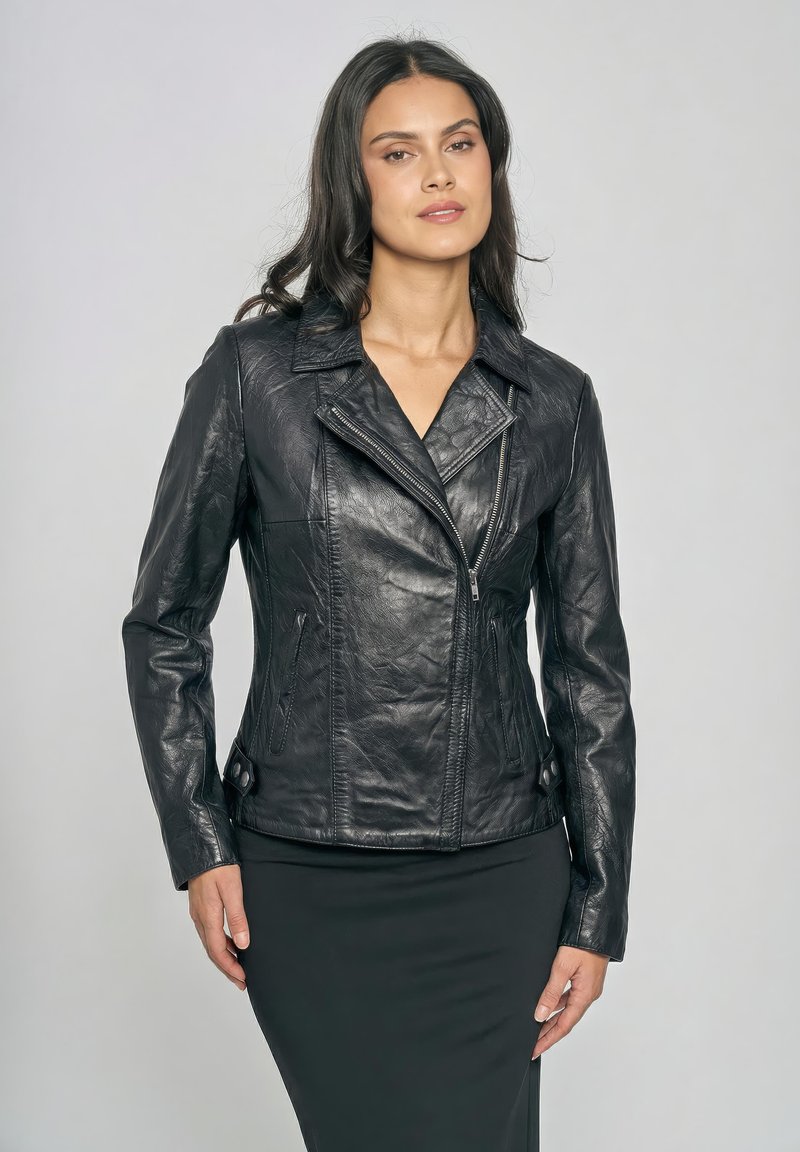Kobieta o długich, ciemnych włosach, ubrana w czarną, teksturowaną skórzaną kurtkę zapiętą do połowy oraz czarną spódnicę, stojąca na jednolitym jasnym tle.