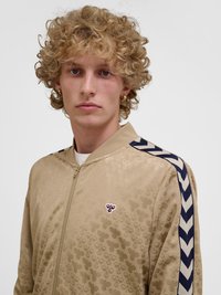 Mlada oseba z kodrastimi blond lasmi, oblečena v bež jakno z zadrgo z ponavljajočimi se vzorci čebel in mornarsko modrimi Chevron črtami na rokavih.