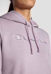 Hoodie lilas en tissu doux avec un logo "Champion" brodé en blanc, doté d'une capuche à cordon et d'une surface texturée.