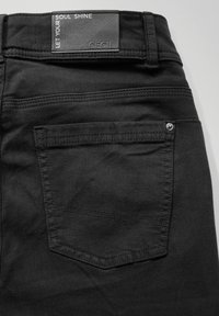Pantalones de mezclilla negros con un bolsillo trasero con una costura vertical y remache de metal, además de una etiqueta cosida en la cinturilla.